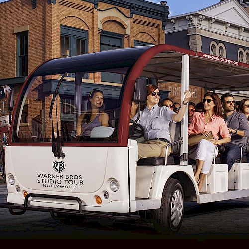 A group of people is sitting on a Warner Bros. Studio Tour tram, enjoying the tour, with a backdrop of studio buildings.