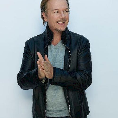 A smiling man claps his hands while standing against a plain background, wearing a black leather jacket and jeans.