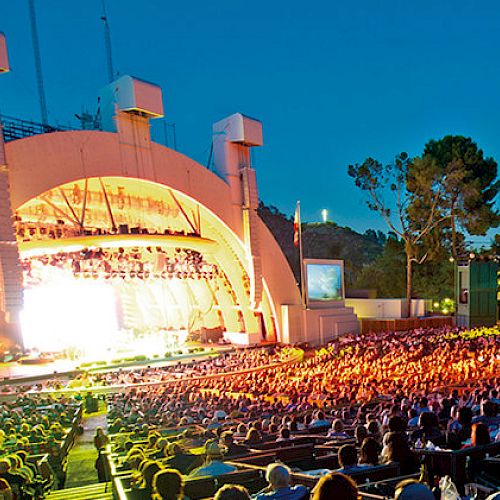 This image shows an outdoor concert at an amphitheater with a large audience under a clear evening sky, lit by vibrant stage lights and surrounded by trees.