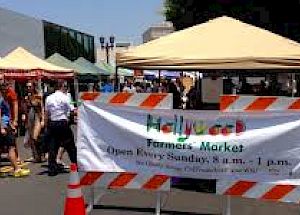 A farmers market with colorful tents and a banner: "Hollywood Farmers' Market, Open Every Sunday, 8 a.m. - 1 p.m." ending the sentence.