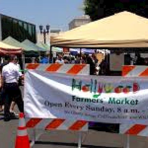 A farmers market with colorful tents and a banner: "Hollywood Farmers' Market, Open Every Sunday, 8 a.m. - 1 p.m." ending the sentence.