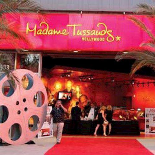 The image shows the entrance of Madame Tussauds Hollywood with a red carpet, palm trees, and large film reel decorations.