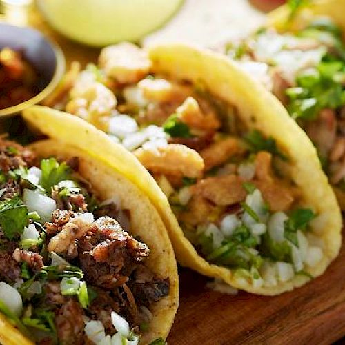 The image shows three tacos on a wooden board, filled with different types of meat, garnished with onions and cilantro, with a side of salsa.