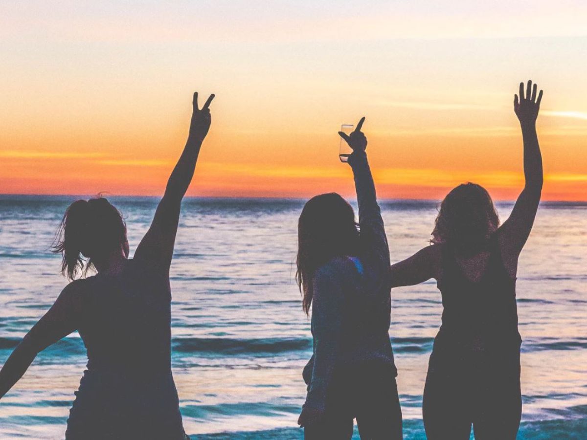 Three people stand at the beach during sunset, raising their arms and making peace signs, with the calm ocean in the background.