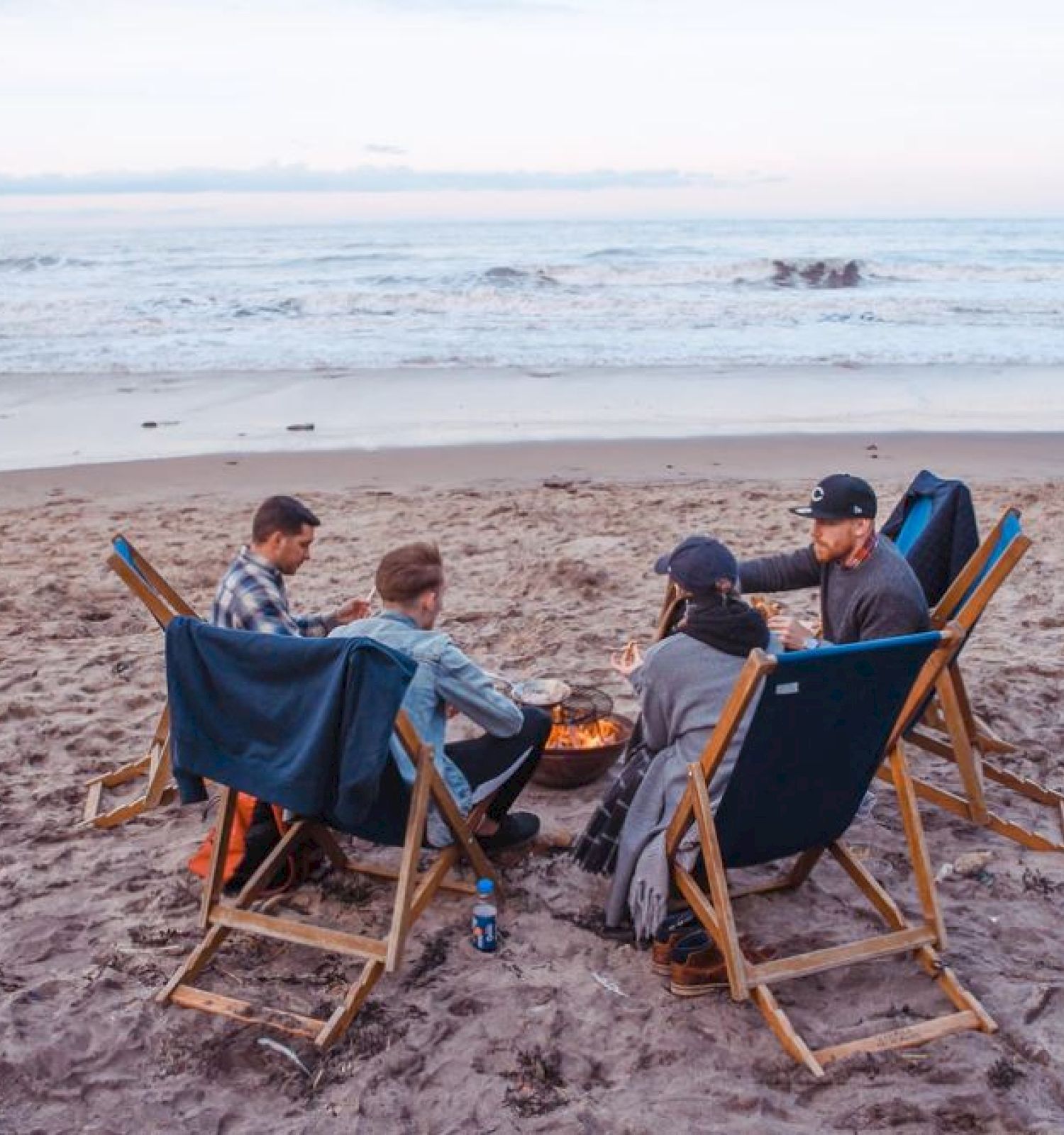 Four people sit in beach chairs around a small fire on a sandy beach, with the ocean in the background as the sun sets.