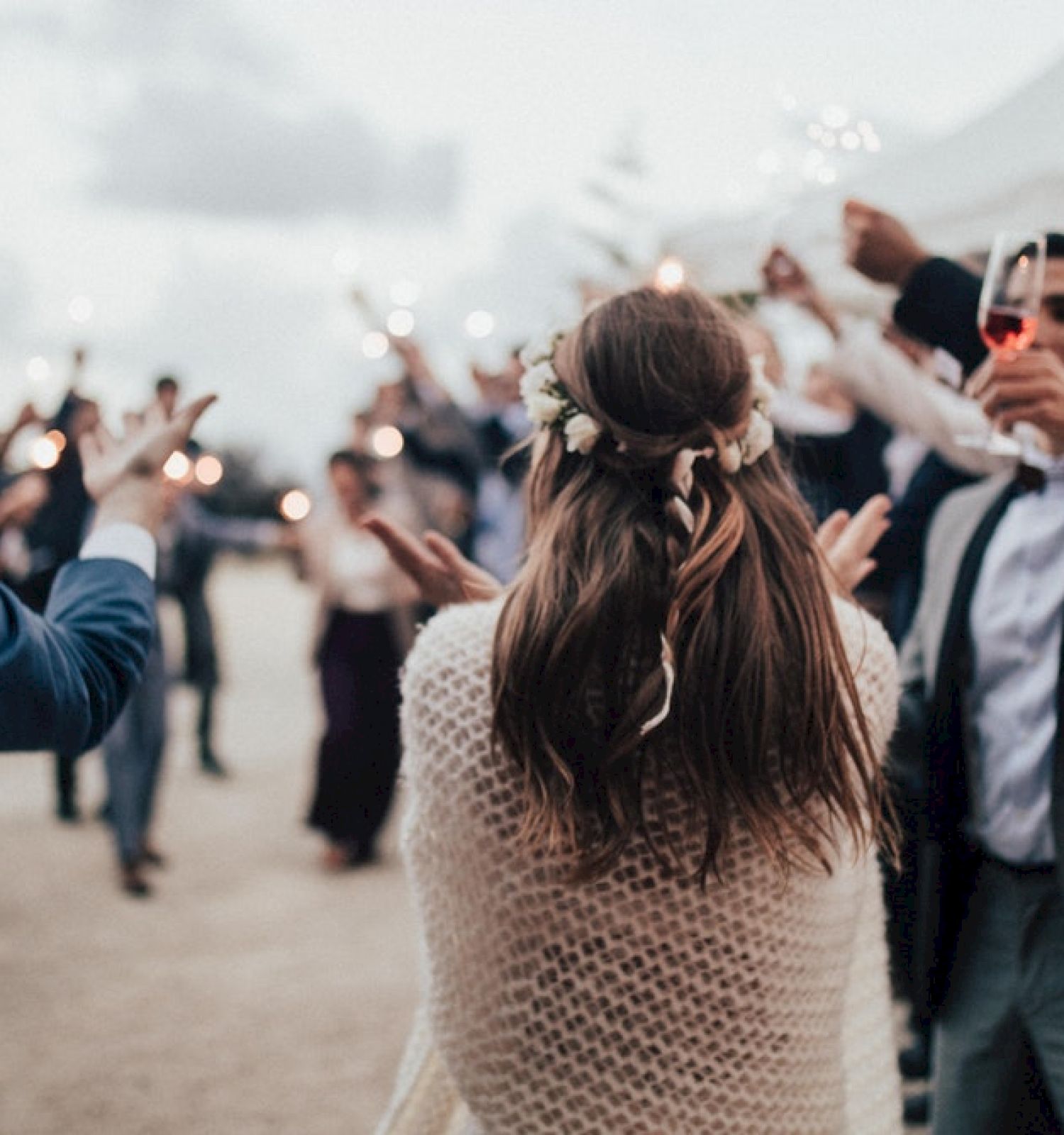 A crowd celebrates with sparklers while a woman in the center with flowers in her hair faces away from the camera, likely at an outdoor event.