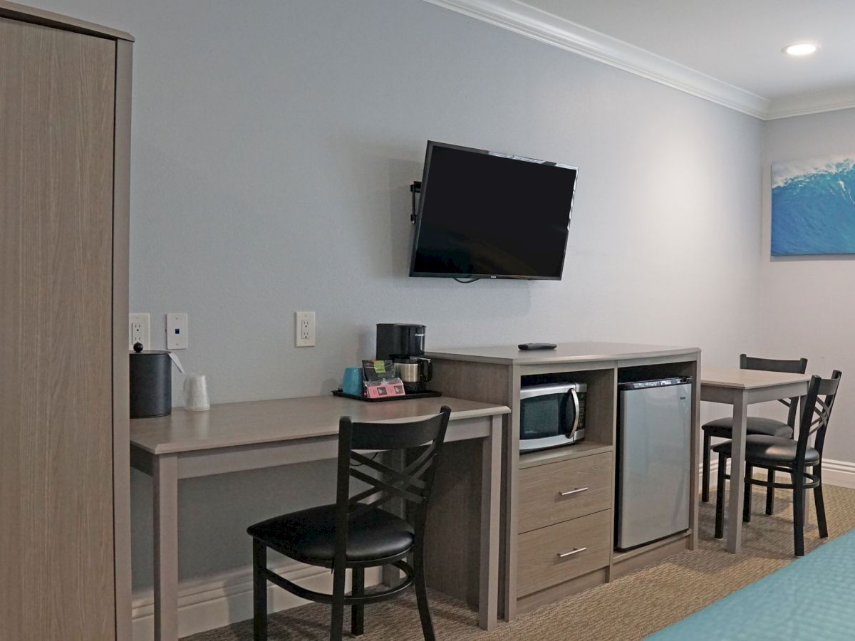 A hotel room features a wardrobe, desk, chairs, wall-mounted TV, microwave, fridge, and artwork of a wave.