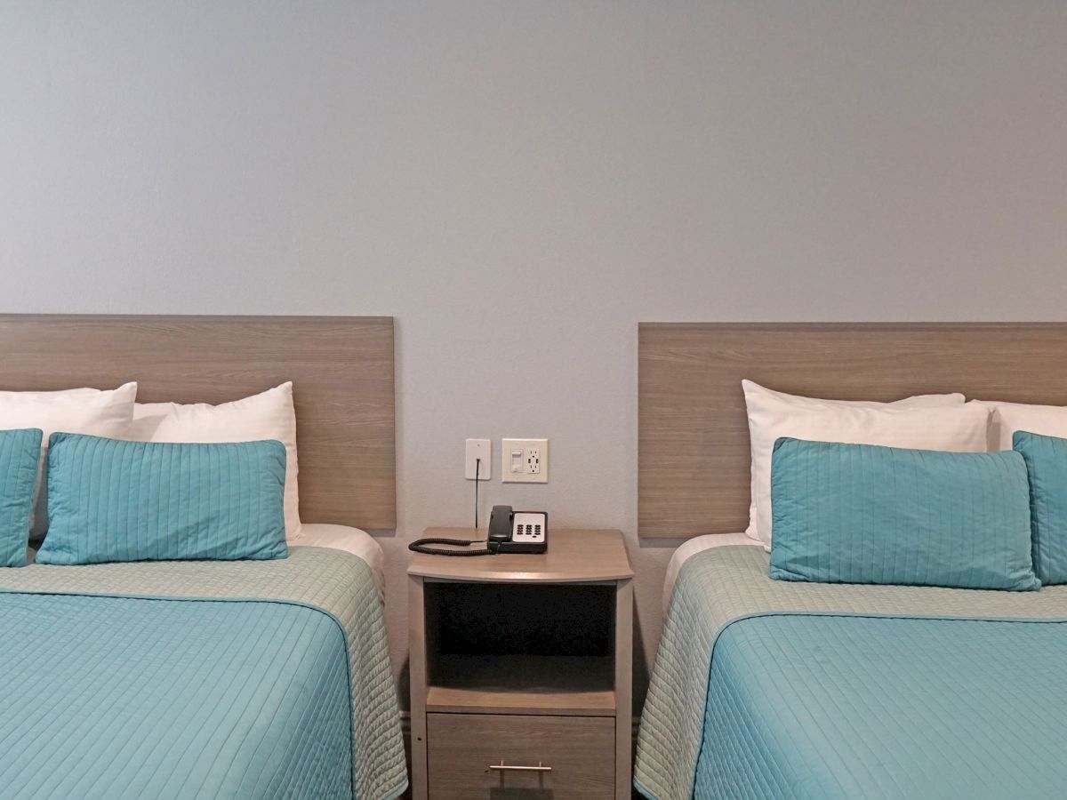 The image shows two beds with blue bedspreads and pillows, separated by a nightstand with a digital clock and a phone on it.