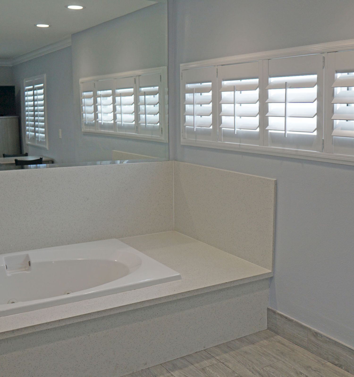 The image shows a modern bathroom with a bathtub, white shutters on the window, and a light grey color scheme, creating a clean, minimal look.