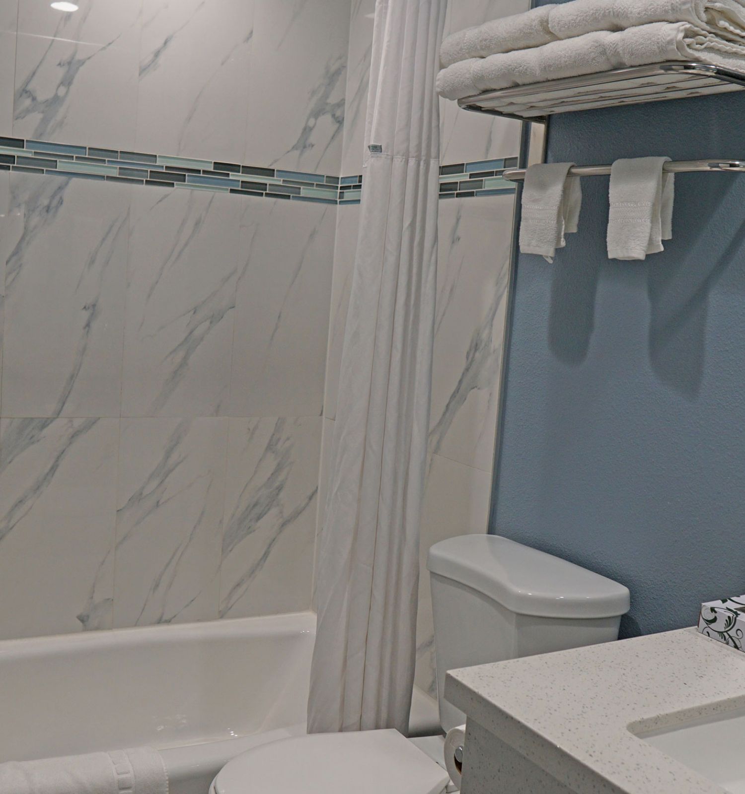 The image depicts a modern bathroom with a white bathtub, toilet, sink, mirror, and towels neatly stacked on a shelf.