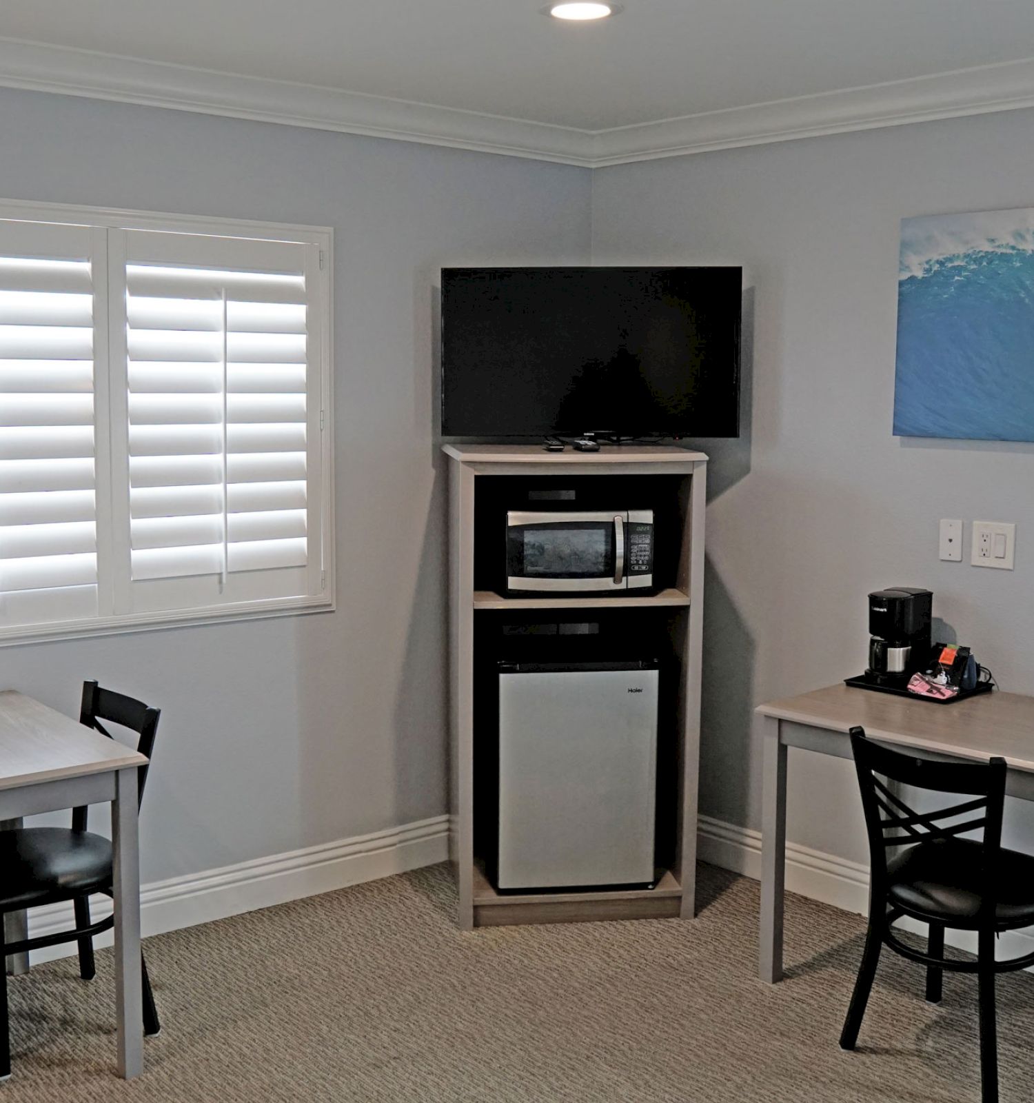 The image shows a room with two small tables and chairs, a mini-fridge, a microwave, a coffee maker, a painting of a wave, and a window with blinds.