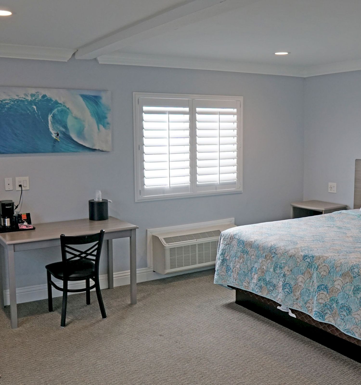 A modern bedroom with a bed, desk, TV, and kitchenette, featuring a beach-themed wall art and a window with white shutters.