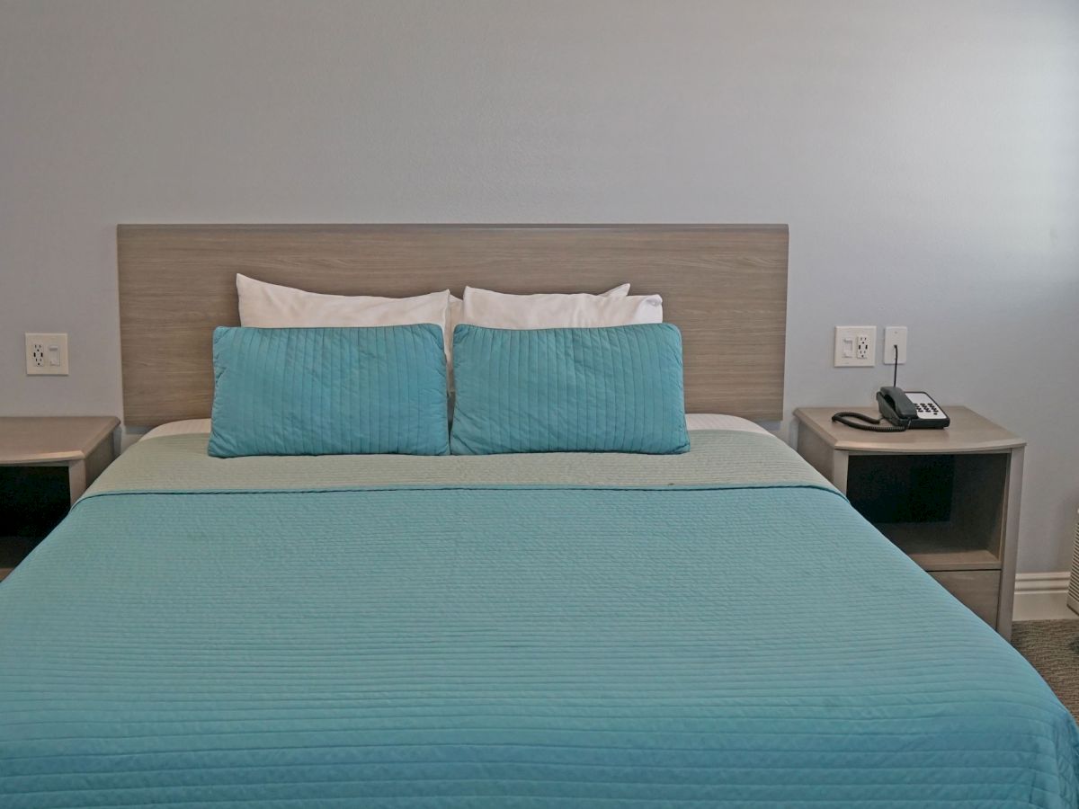 The image shows a neatly made bed with blue bedding and pillows, flanked by two nightstands, one with a telephone and the other empty.