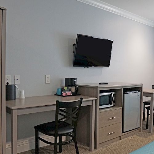 The image shows a hotel room with a desk, chairs, a mini-fridge, microwave, wall-mounted TV, and a picture of a wave on the wall.