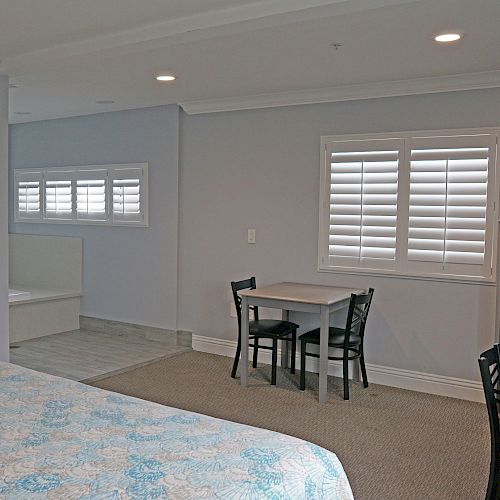 The image shows a room with a bed, a small dining table with two chairs, a wall-mounted TV, a microwave, and windows with shutters.