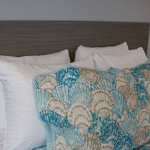 A neatly made bed with white pillows and a headboard, featuring bedding with a seashell pattern, next to a nightstand with a black telephone.