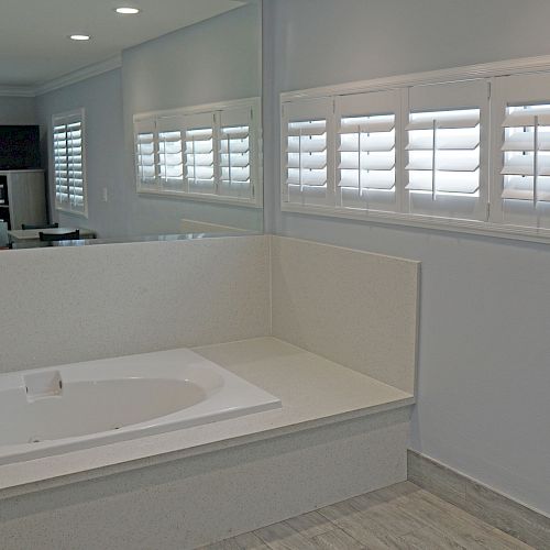 A modern bathroom featuring a large bathtub, white shutters on the windows, and a mirror reflecting part of the room.