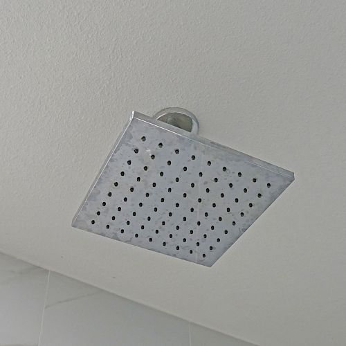 The image shows a square, metal showerhead attached to the ceiling, designed to disperse water through multiple small holes, commonly used in bathrooms.