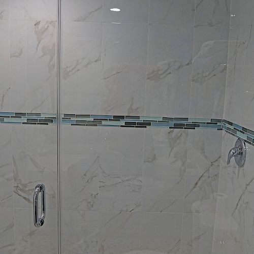 A modern shower with white marble walls, a glass door, a rain shower head, and a decorative horizontal tile strip, featuring a tiled mosaic floor.