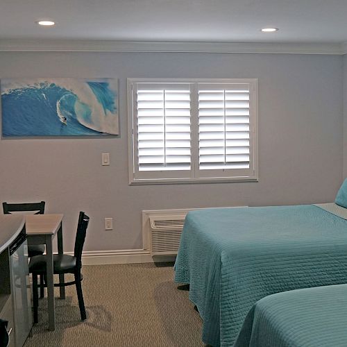 The image shows a neatly arranged hotel room with two beds, a wall-mounted TV, a small desk and chair, and a picture of waves on the wall.