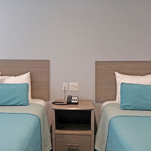 The image shows two neatly made beds with light blue bedding, separated by a nightstand with a telephone and a small digital clock, ending the sentence.