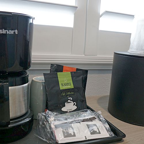 The image shows a coffee maker, coffee packets, and an ice bucket on a tray on a countertop near a window with blinds ending the sentence.