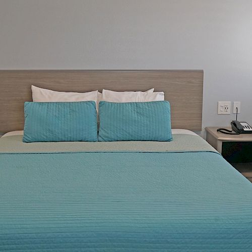 This image shows a neatly made bed with blue bedding, two pillows, and nightstands on each side. There is a phone on the right nightstand.