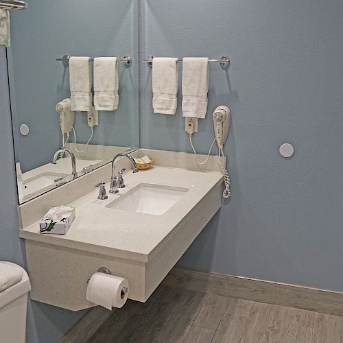 A bathroom with a sink, large mirror, towels, a wall-mounted hairdryer, toilet, and a door. The walls are blue and the floor is wooden.