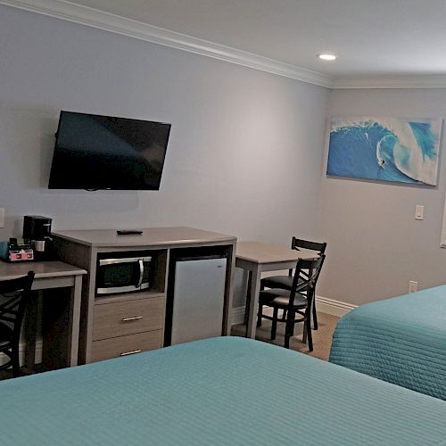 A hotel room with two beds, a wall-mounted TV, a fridge, a microwave, a desk with chairs, and a window with shutters, decorated with a wave painting.