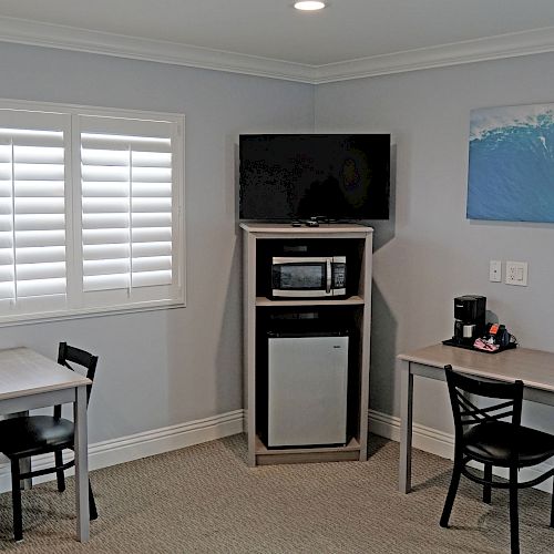 A room with two tables and chairs, a small refrigerator, microwave, coffee maker, a window with blinds, and a painting of a wave on the wall.