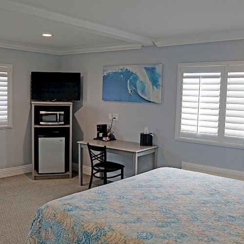 A cozy room features a bed, TV, microwave, mini fridge, small table with chairs, beach-themed artwork, and shuttered windows, creating a serene ambiance.