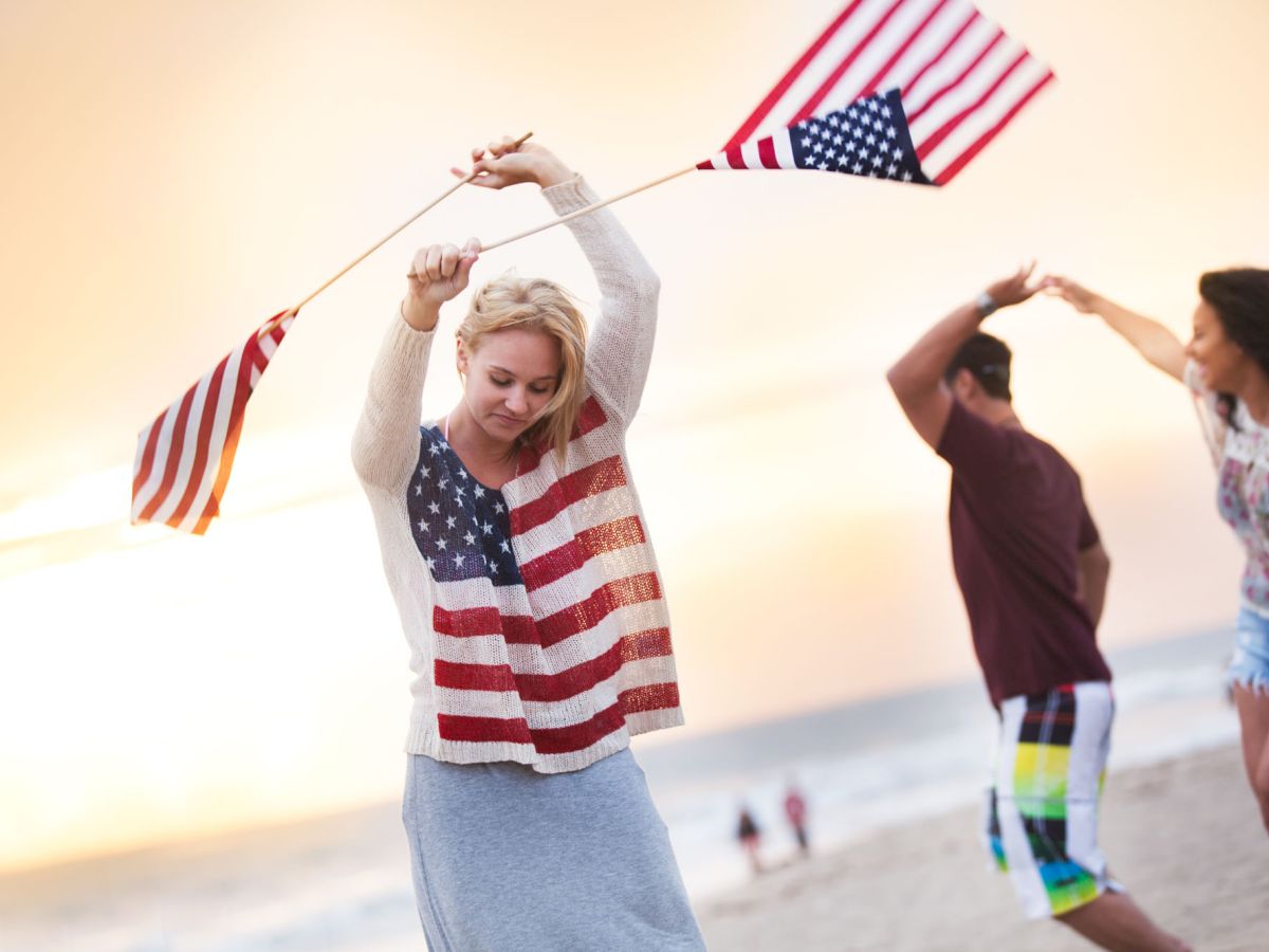 Three people are waving flags and dancing on a beach at sunset. The two women have American flag-themed clothing, and a man is twirling another woman.