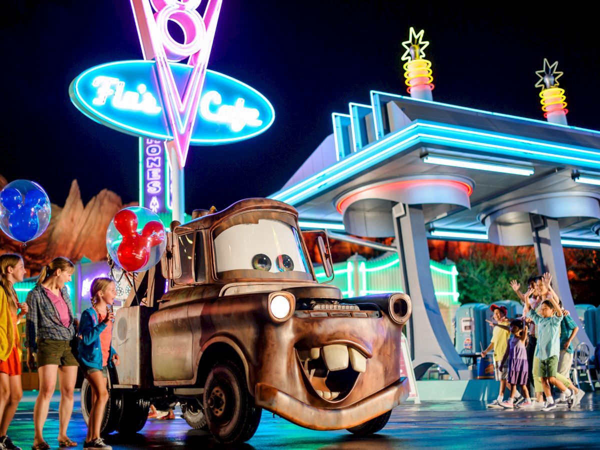 The image shows people enjoying themselves near a themed café with a car character that has a smiling face at night.