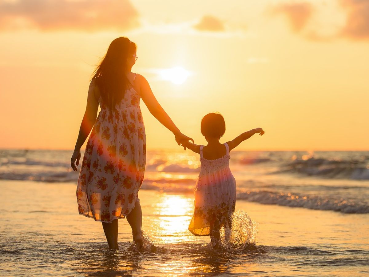 A woman and child, both in dresses, hold hands and walk along the shore during a beautiful sunset, enjoying the beach and the gentle waves.