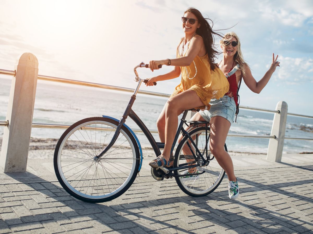 Two women ride a bicycle along a promenade by the sea, one pedaling and the other enjoying the ride while making a peace sign.