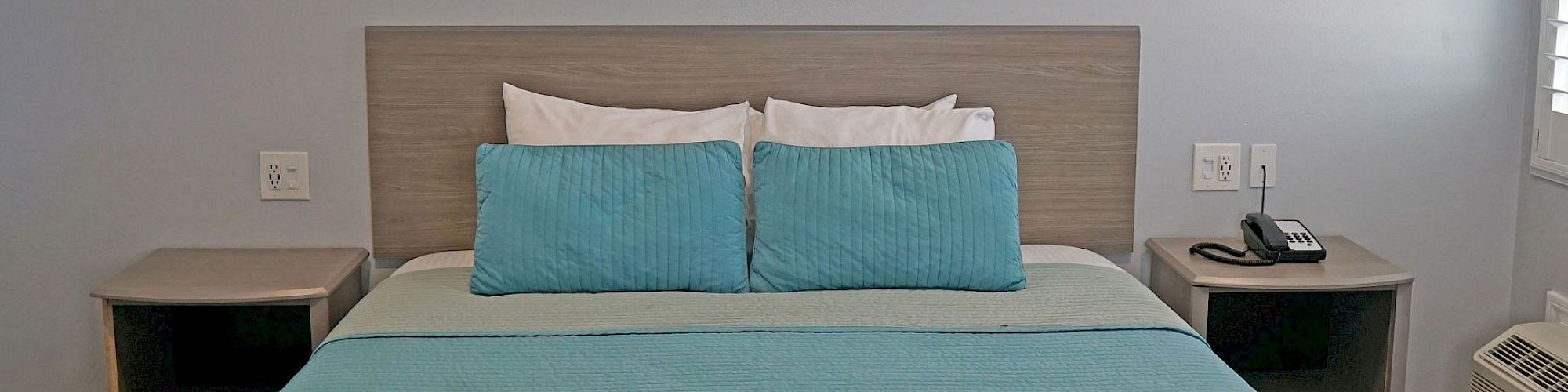 A neatly made bed with turquoise bedding, two teal pillows, and two nightstands with lamps and a phone, set in a simple room.