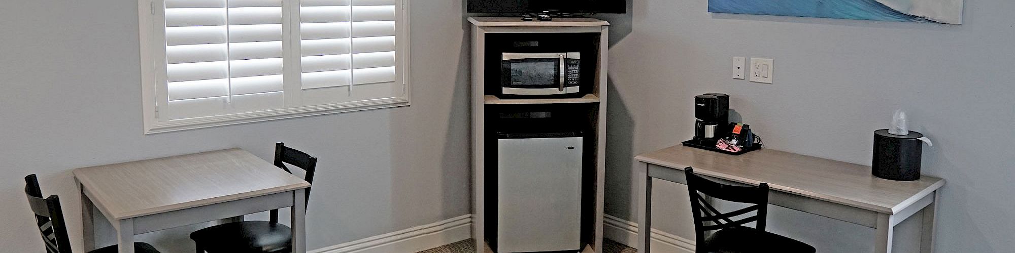 The image shows a small room with two tables and chairs, a TV, a microwave, a mini-fridge, a coffee maker, and a wave painting on the wall.