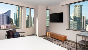 A modern hotel room has large cityscape windows, a bed, a TV on the wall, a chair, and a table.