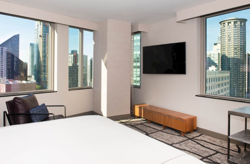 A modern hotel room has large cityscape windows, a bed, a TV on the wall, a chair, and a table.