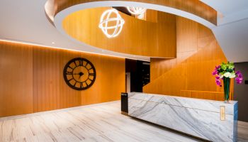 A modern lobby area with a marble reception desk, wooden wall panels, a large clock, a spiral staircase, and hanging light fixtures.