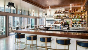 A modern bar with a wooden counter, stylish bar stools, and a well-stocked shelf of various alcoholic beverages in a well-lit, open space.