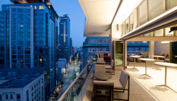 A modern cityscape view from a high-rise balcony at dusk, with a mix of residential and commercial buildings, and outdoor seating and tables.
