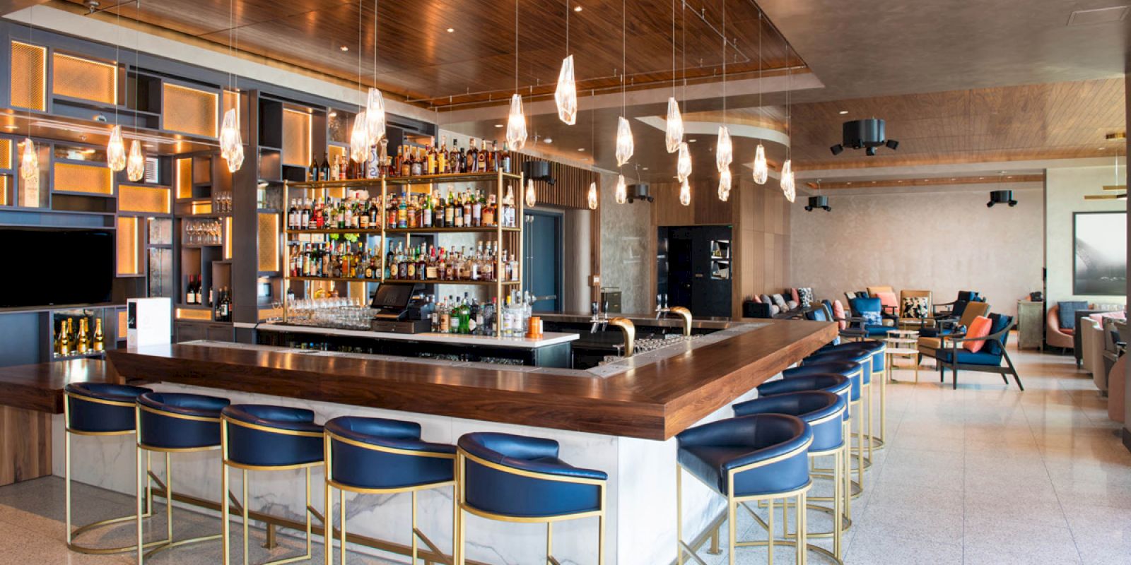 A modern bar with blue cushioned stools, hanging lights, a variety of bottles on the shelves, and a seating area in the background.