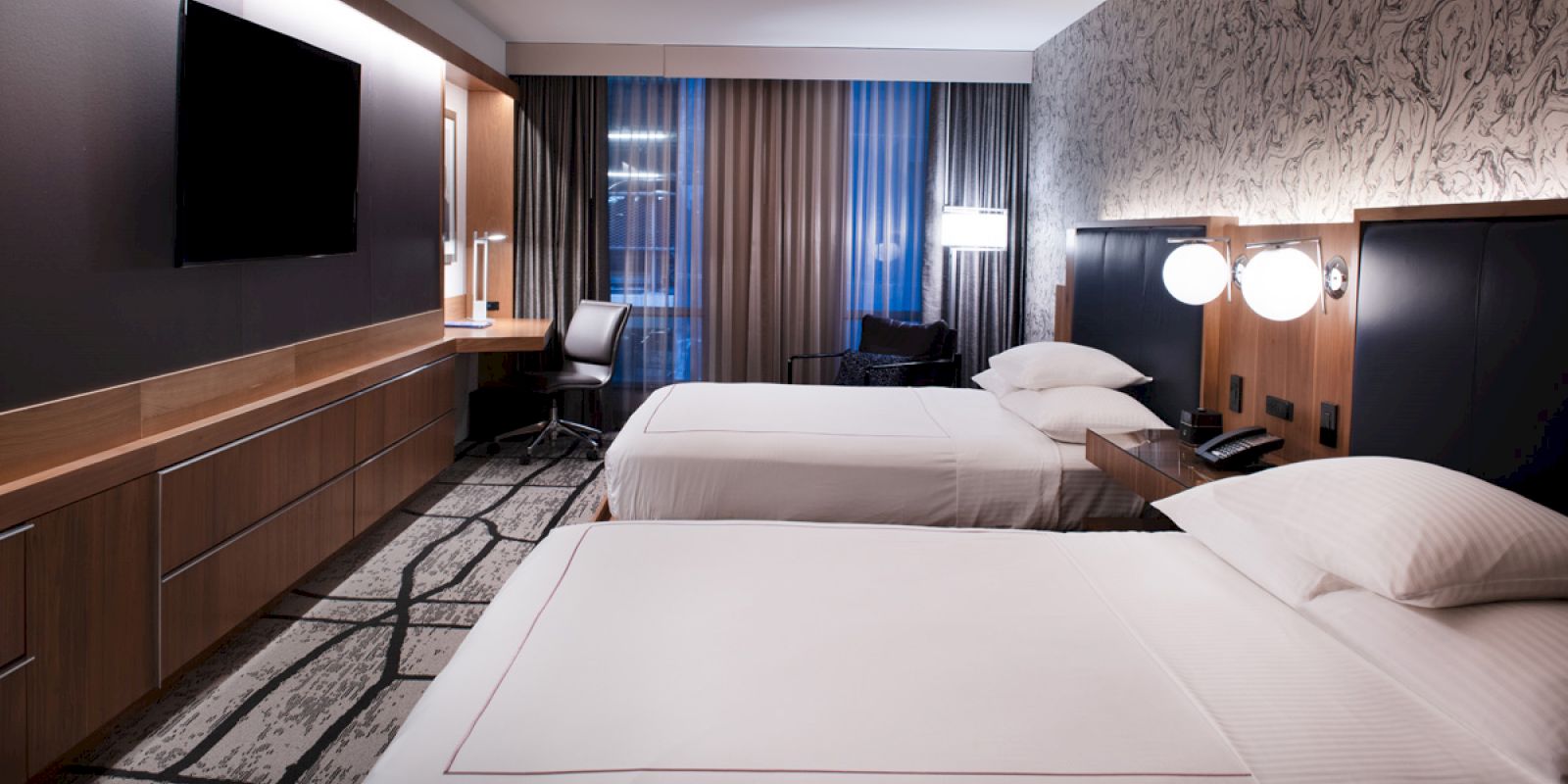 A modern hotel room with two beds, a TV, a desk with a chair, and a window with curtains. The room features stylish decor and ample lighting.