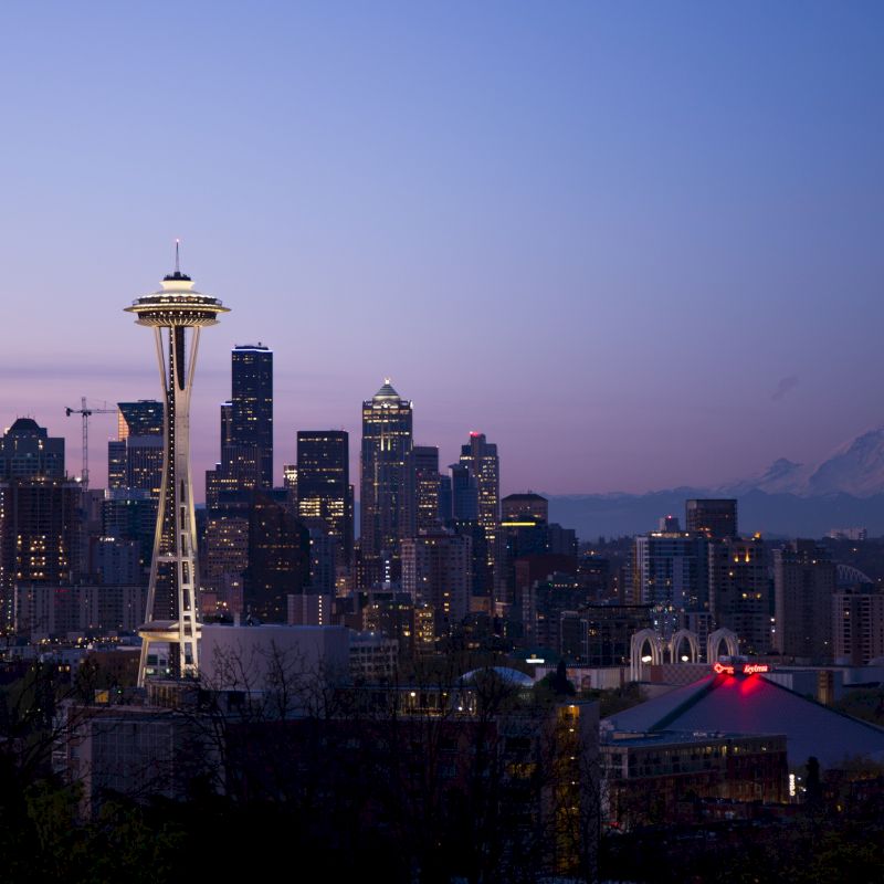 The image depicts a city skyline at dusk with the Space Needle prominently featured, surrounded by buildings, and a mountain in the background.