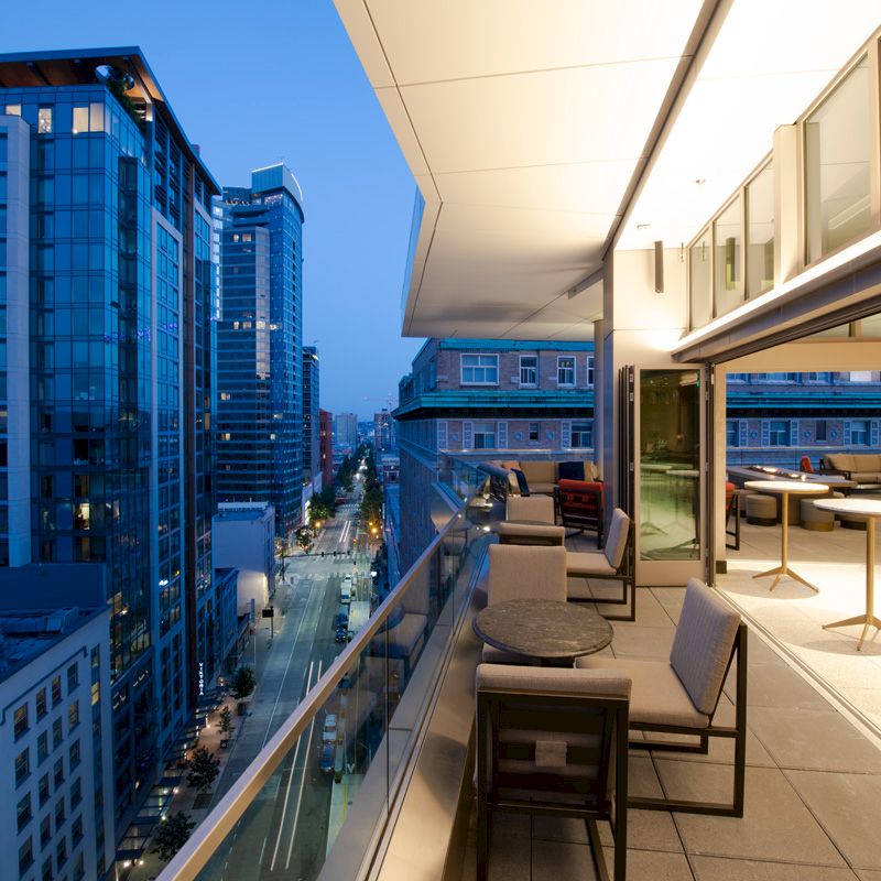 A modern cityscape at dusk, featuring tall buildings and streets alongside a sleek outdoor balcony with furniture and tables.