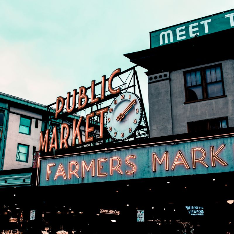 The image shows a public market sign with a clock above it and a larger "Farmers Market" sign below, set against a backdrop of buildings.