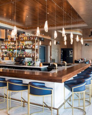 A modern bar with a wooden counter, blue chairs, hanging lights, and a fully stocked shelf of various alcohol bottles; seating area in the back.