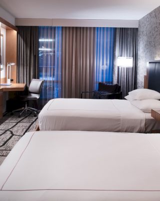 A modern hotel room with two beds, a TV, a desk, and floor-to-ceiling windows with curtains, featuring contemporary decor and ambient lighting.