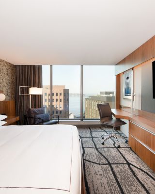 A modern hotel room with a large bed, wall-mounted TV, desk, chair, and floor-to-ceiling windows offering a city view and partial water view.